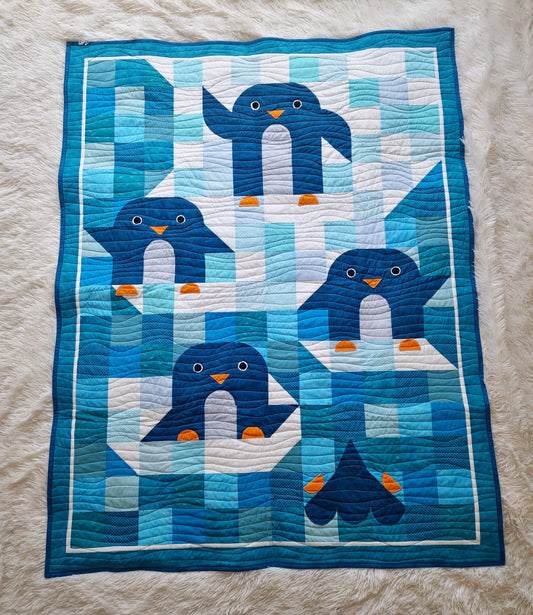 Top down view of the Chill and Thrill toddler penguin quilt laid flat showing four penguins in scrappy blue and teal patchwork with wave quilting and dark blue border