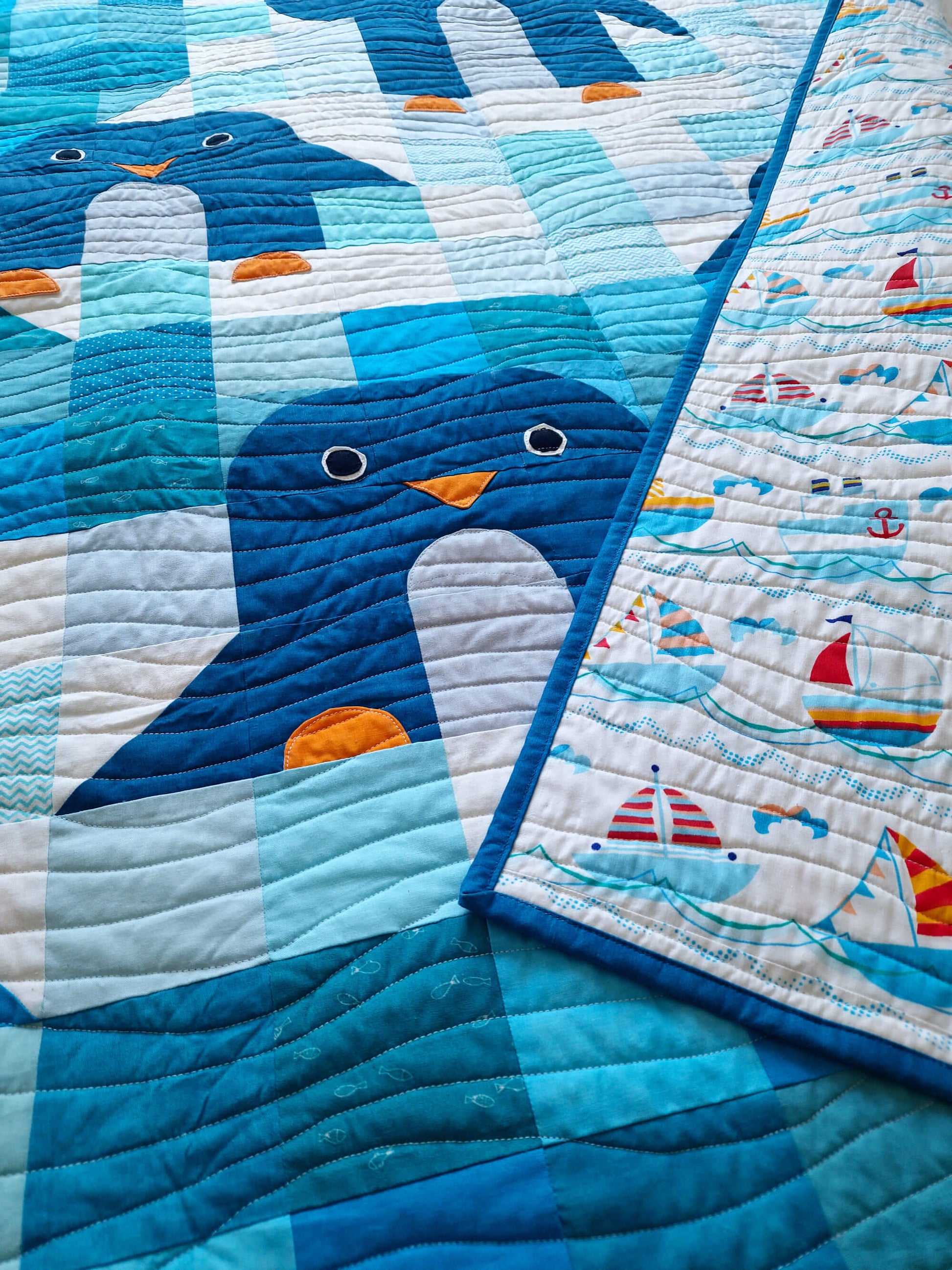 Close up of a penguin on the Chill and Thrill baby quilt showing the navy body, white belly, orange beak and feet, with the nautical sailboat print backing fabric visible alongside
