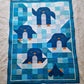 Top down view of the Chill and Thrill toddler penguin quilt laid flat showing four penguins in scrappy blue and teal patchwork with wave quilting and dark blue border