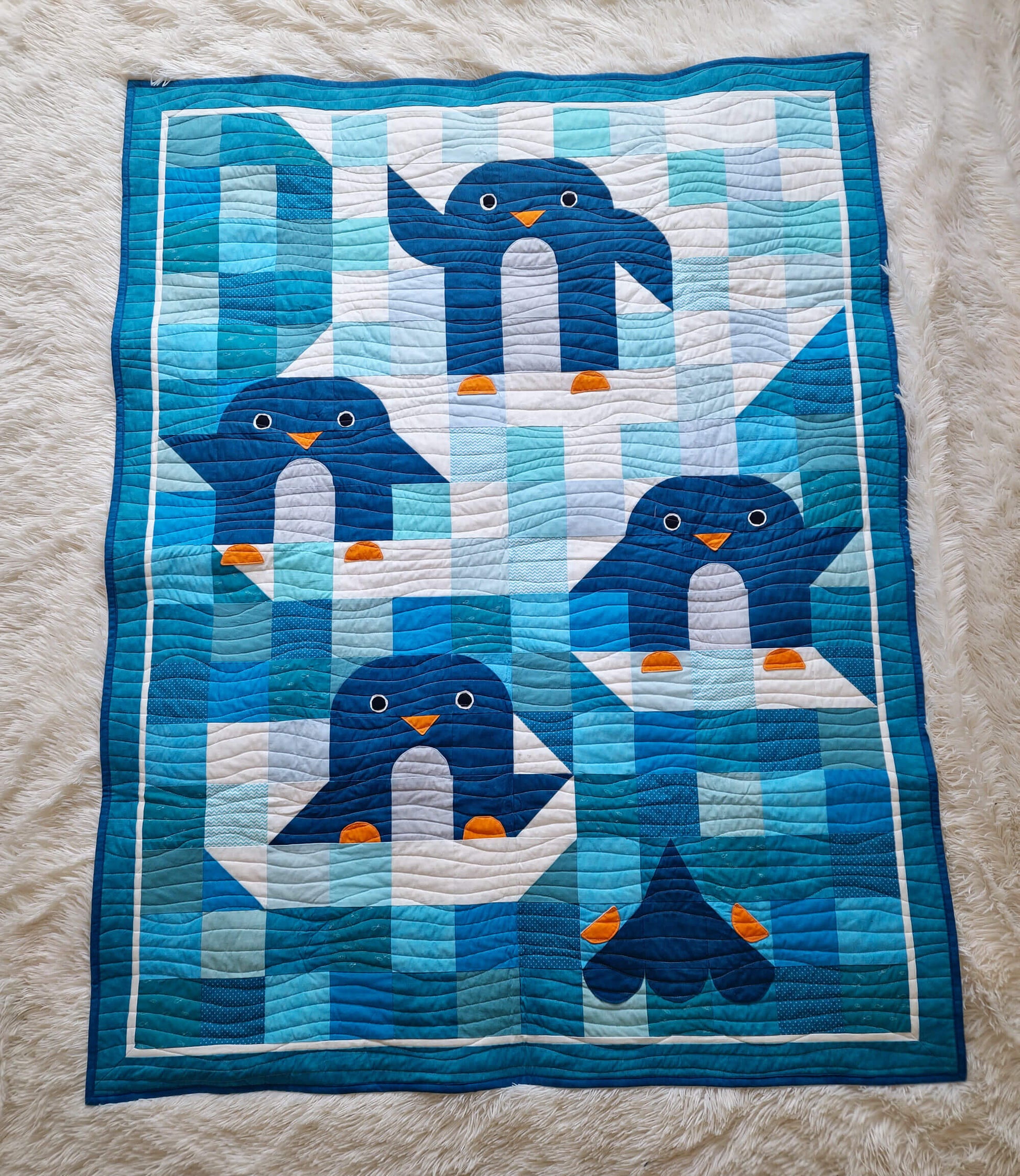 Top down view of the Chill and Thrill toddler penguin quilt laid flat showing four penguins in scrappy blue and teal patchwork with wave quilting and dark blue border