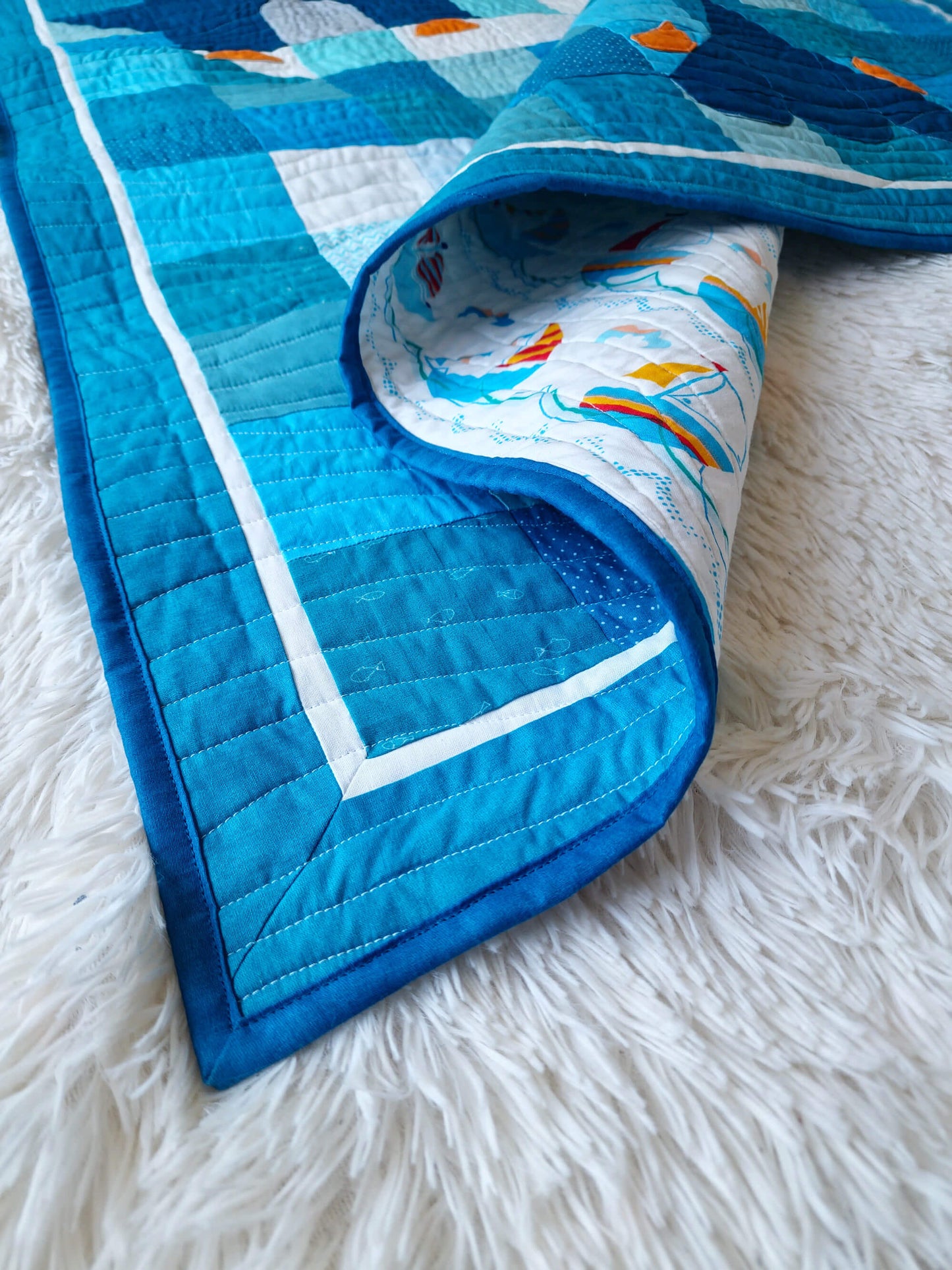 Corner of the Chill and Thrill baby quilt folded back to reveal the nautical sailboat print backing fabric, with dark blue binding and scrappy blue patchwork visible on the front