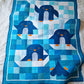 Top down view of the Chill and Thrill toddler penguin quilt laid flat on a white fluffy surface showing four playful penguins in scrappy blue patchwork with orange beaks and feet