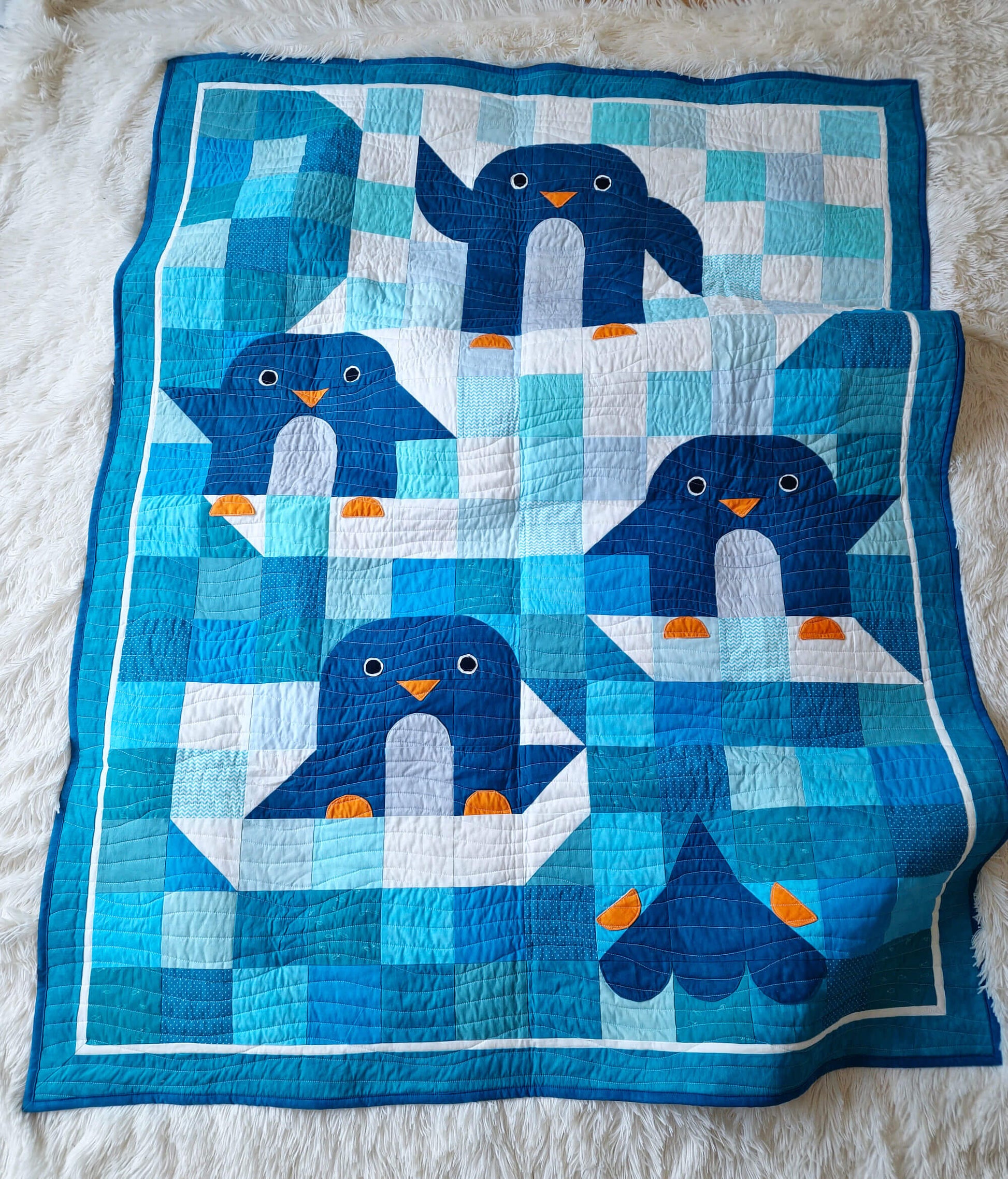 Top down view of the Chill and Thrill toddler penguin quilt laid flat on a white fluffy surface showing four playful penguins in scrappy blue patchwork with orange beaks and feet