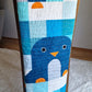 Chill and Thrill penguin baby quilt draped over a wooden chair showing the full length of the quilt with a close up penguin appliqué featuring orange beak and white belly