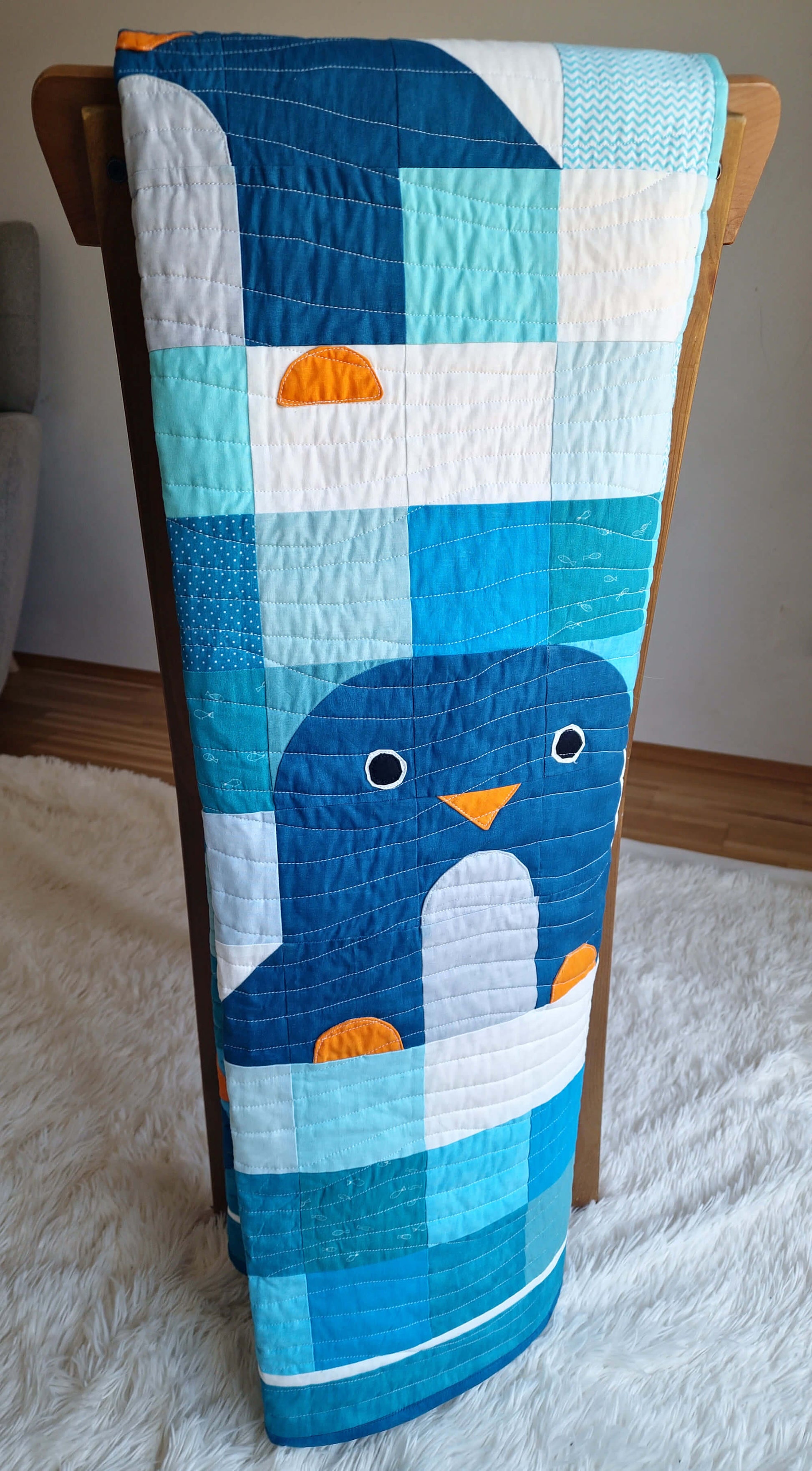 Chill and Thrill penguin baby quilt draped over a wooden chair showing the full length of the quilt with a close up penguin appliqué featuring orange beak and white belly