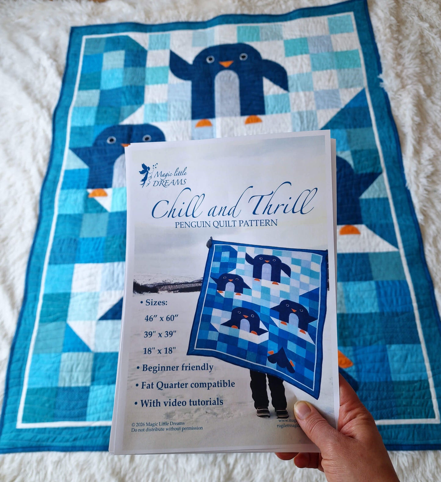 Hand holding the printed Chill and Thrill penguin quilt pattern PDF in front of the finished quilt laid on a white surface showing the pattern cover with Lapland photo
