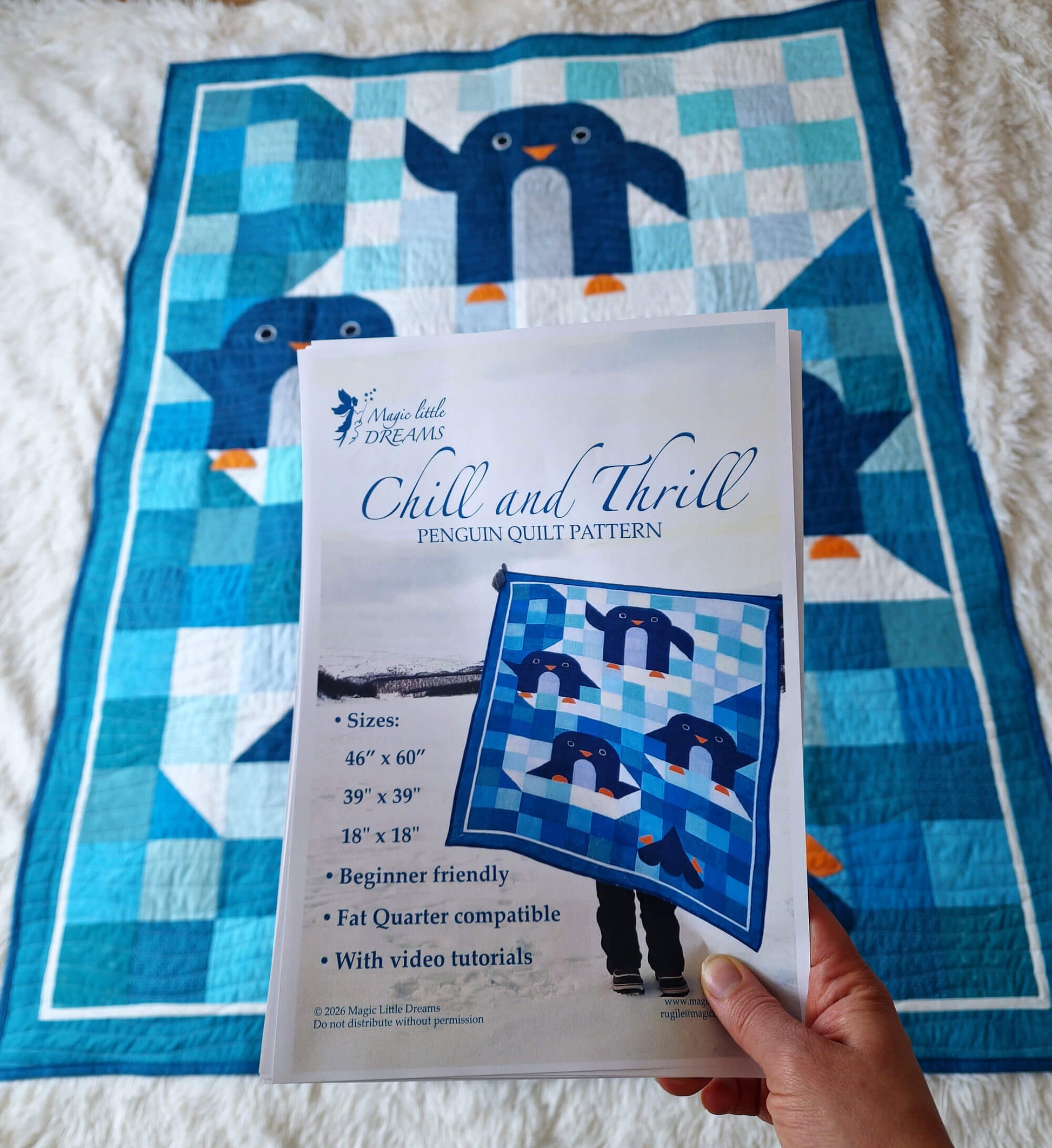 Hand holding the printed Chill and Thrill penguin quilt pattern PDF in front of the finished quilt laid on a white surface showing the pattern cover with Lapland photo
