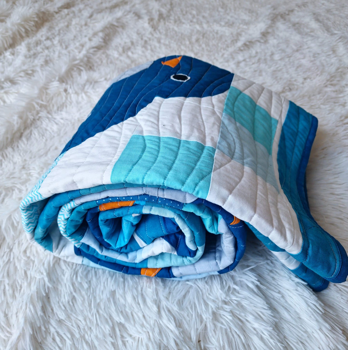 Chill and Thrill baby boy penguin quilt rolled up on a white fluffy surface showing the soft drape and thickness of the quilt with a penguin face and orange beak visible on top