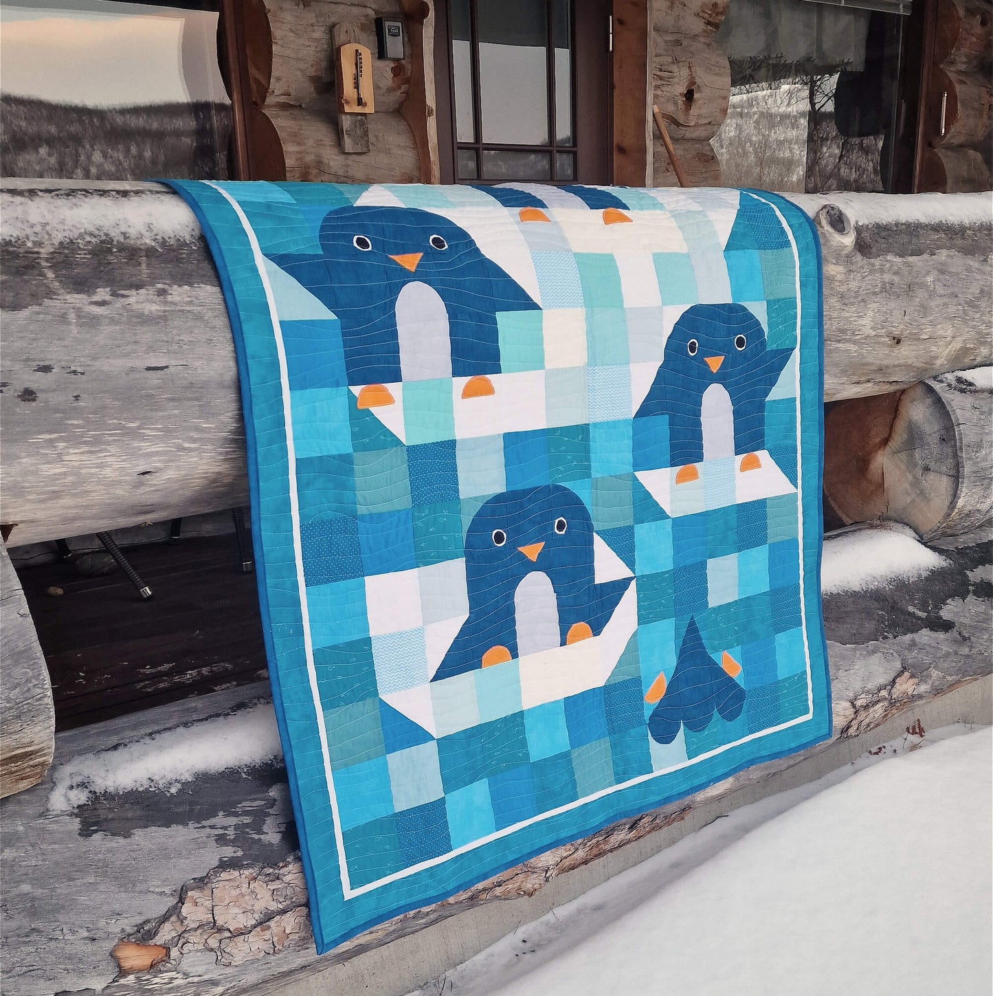 Chill and Thrill penguin baby quilt draped over a snow covered log bench on a cabin porch in Lapland showing the full quilt with playful penguin appliqués in winter setting
