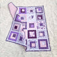 Purple and white quilt with square patterns and baby footrpints on a white fluffy surface