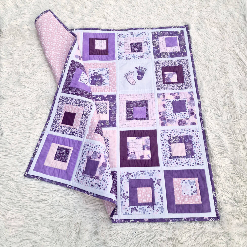 Purple and white quilt with square patterns and baby footrpints on a white fluffy surface